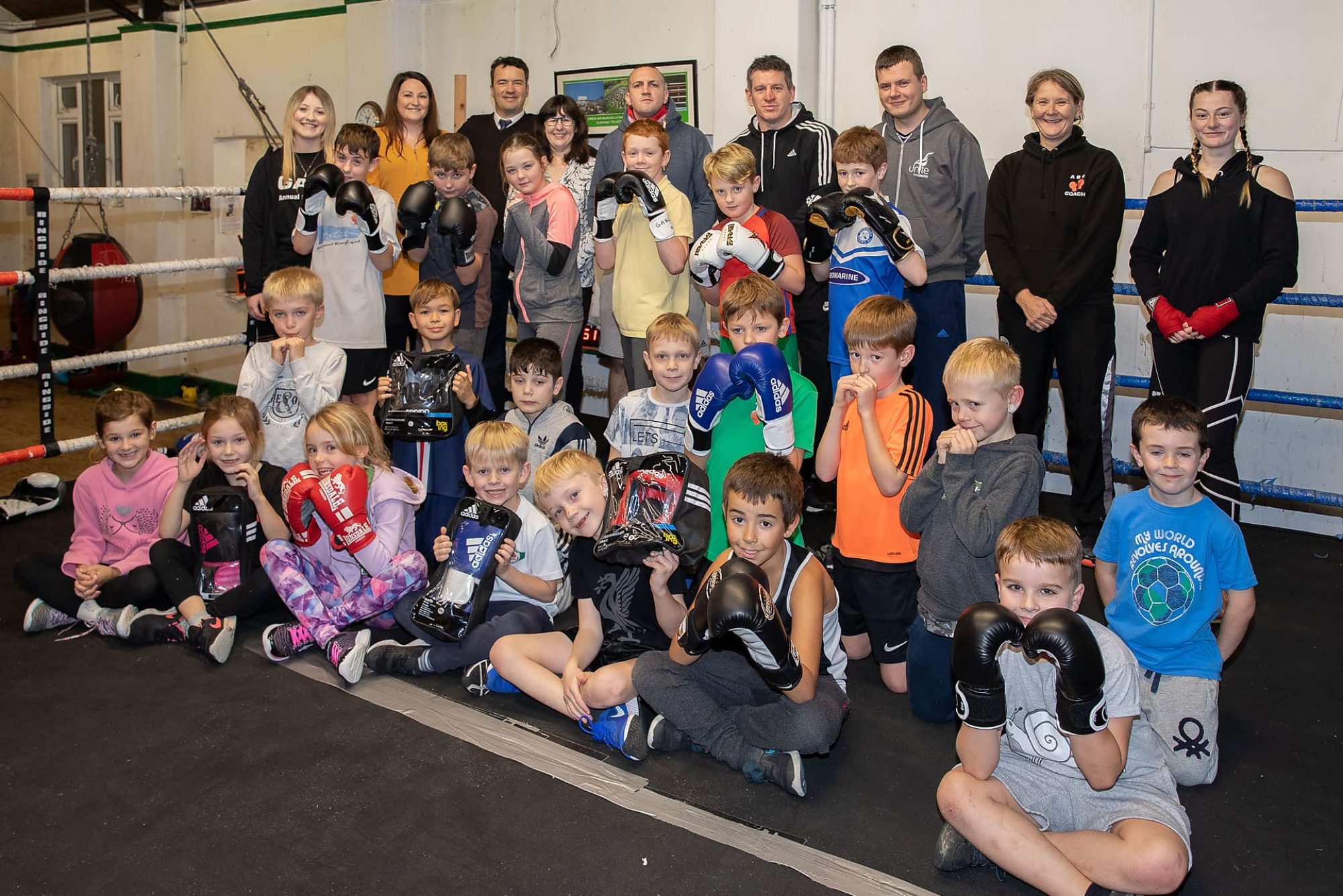 A group of children sitting in a boxing ring with gloves on posing for the camera