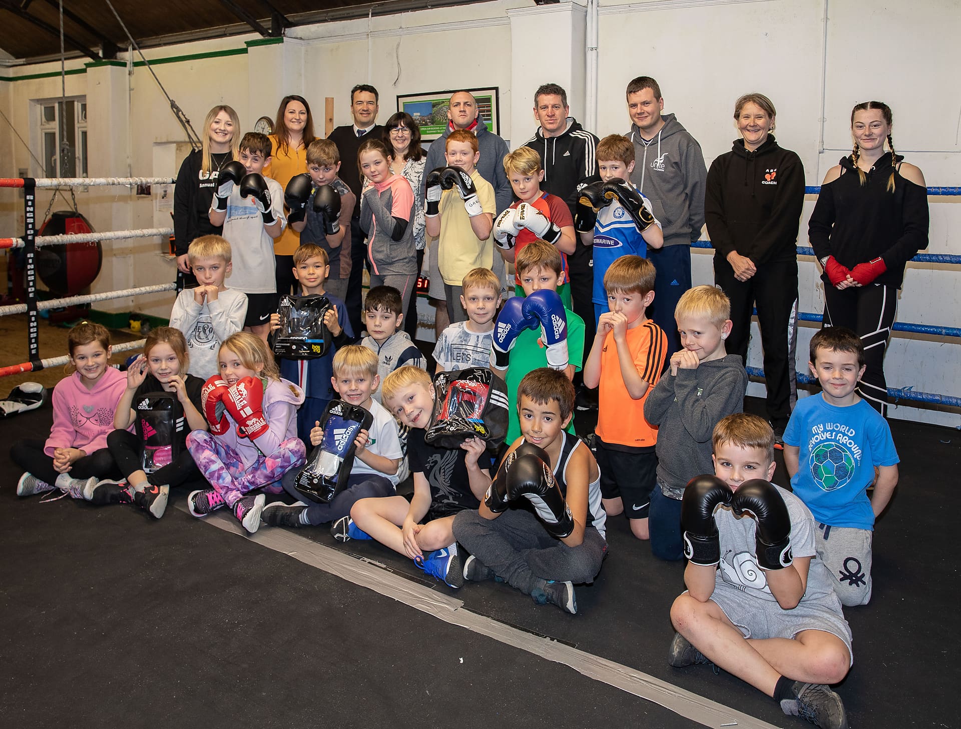 Many children sat in a boxing ring, wearing boxing gloves, smiling for a group photo