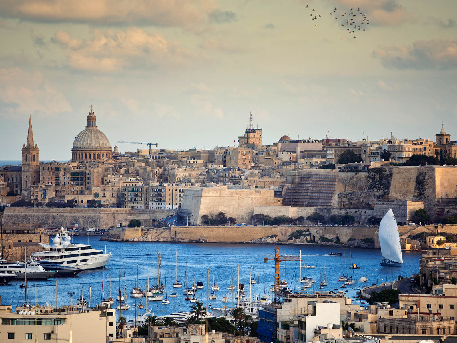 A river with many boats docked. A view of Malta architecture in the background
