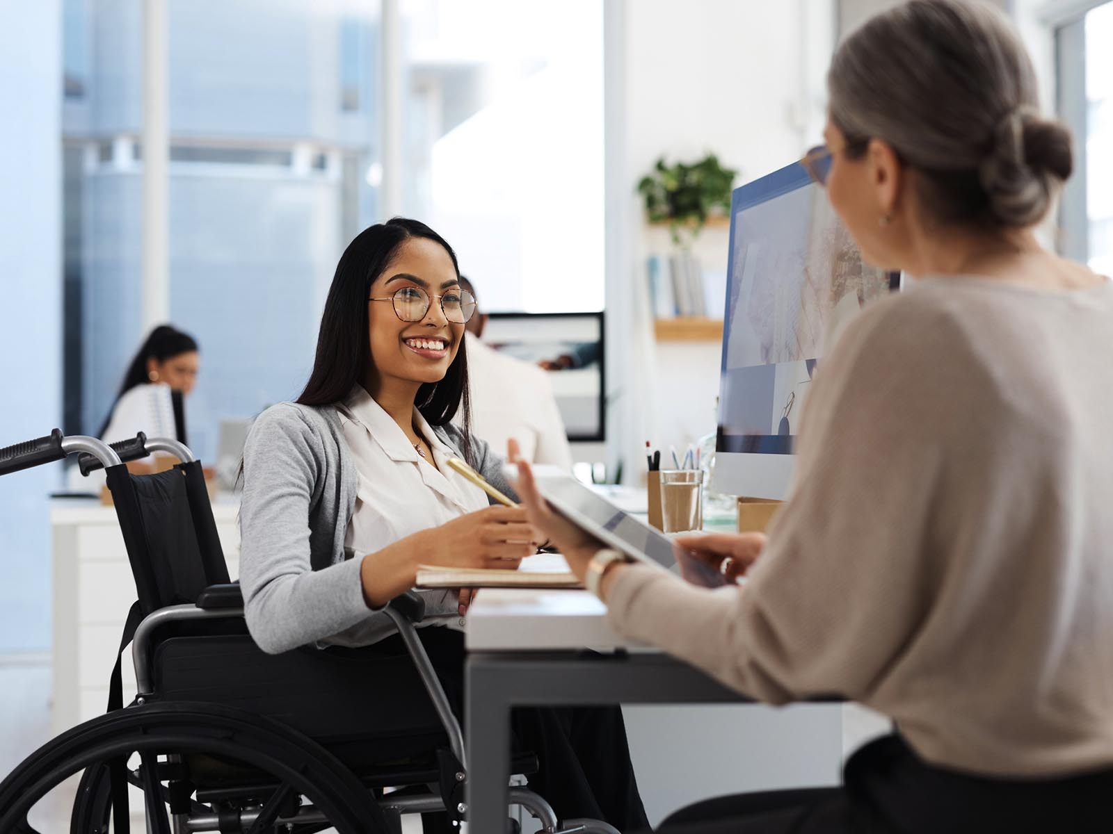 A woman in a wheelchair having a pleasant conversation with another woman while sat at her desk