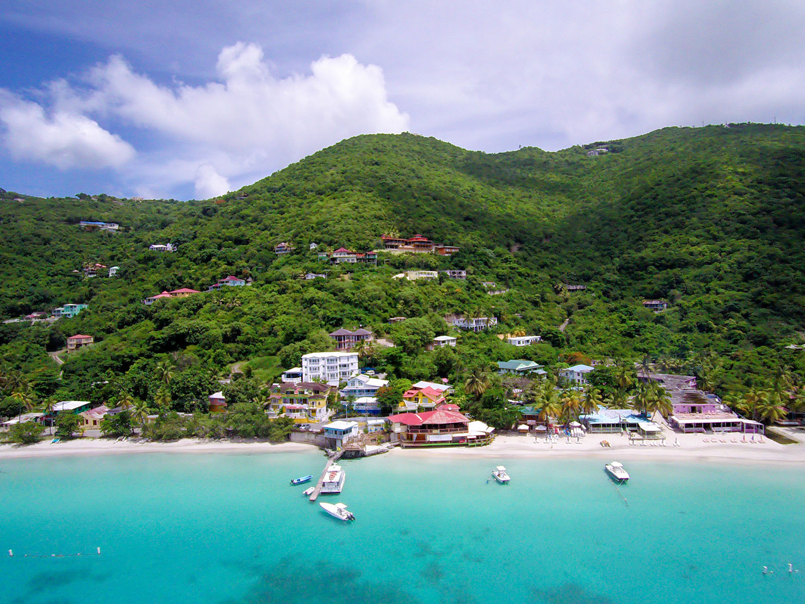 An image of the crystal blue sea with a few boats docked. Green hills in the background with many houses hiding in the shrubbery